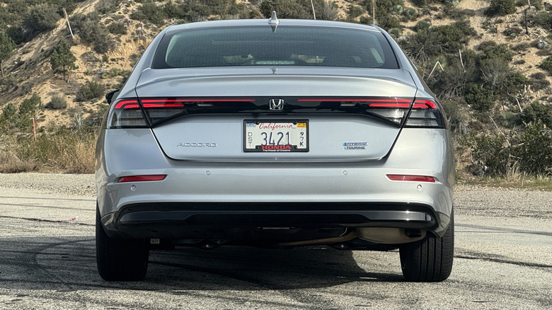 A rear shot of the silver Accord Hybrid Touring parked on asphalt in front of a shrub-covered mountainside