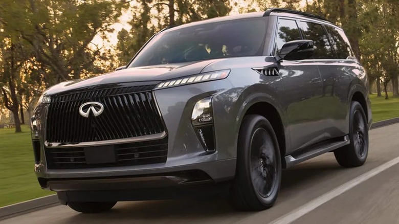 A gray Infiniti QX80 on the road in front of grass and trees