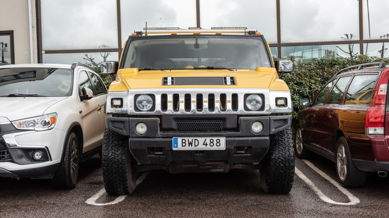 A yellow Hummer H2 parked in a small space, with a white car on its left and a red one on its right, and little space between them