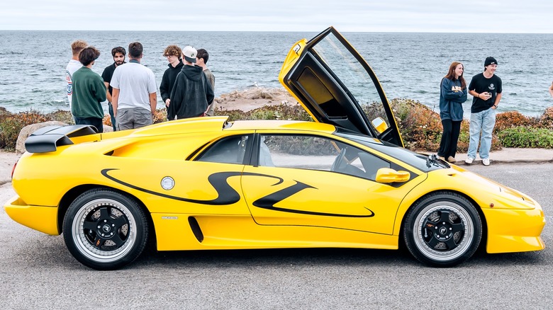A yellow Lamborghini Diablo SV sits on pavement in front of small groups of people, some bushes, and the ocean
