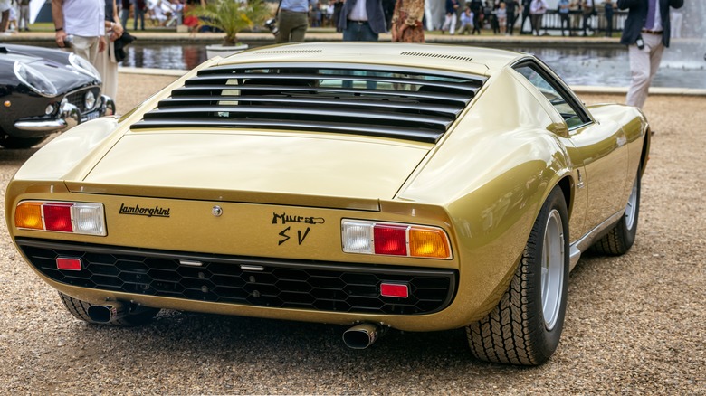 A gold Lamborghini Miura SV sits on sandy, pebbly ground next a black Ferrari 250 in front of a fountain with party guests milling around