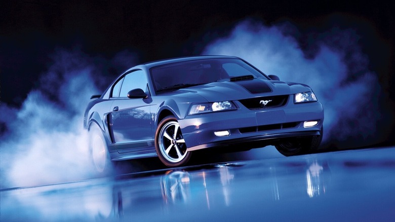 A blue 2003 Mustang Mach 1 with smoke rising from its rear against a black background