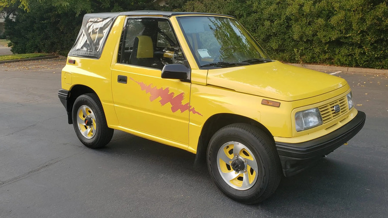 A front three-quarters shot of a yellow Geo Tracker soft top parked on asphalt in front of a hedge