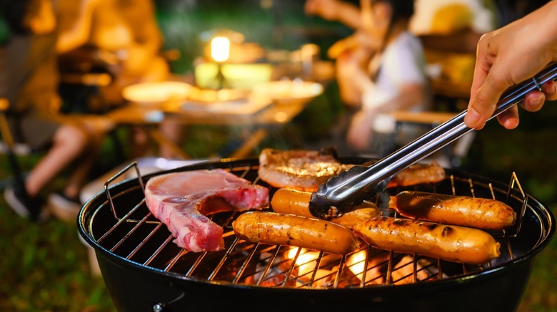 People grilling meats while camping on a small charcoal grill.