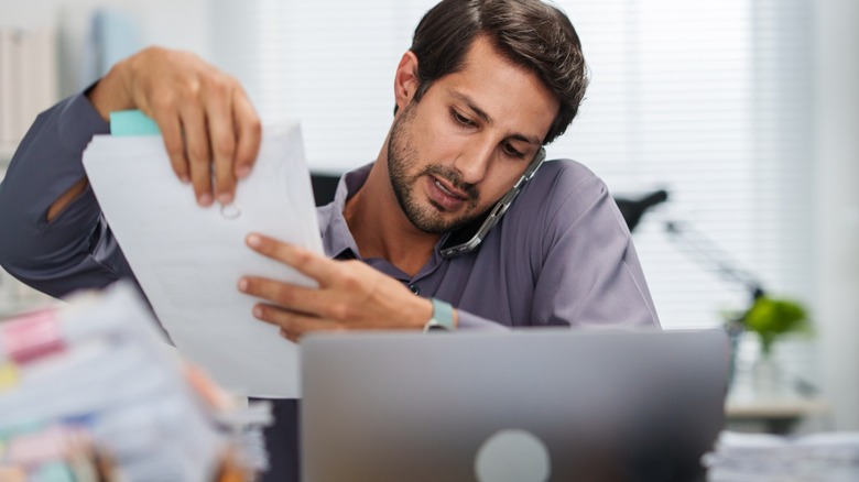A busy man on the phone in front of a laptop, trying to handle a stack of papers
