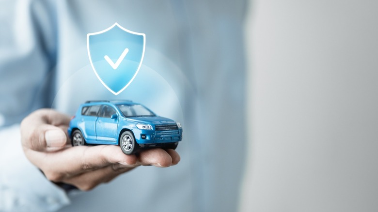 A hand holds a toy car while a blue glowing shield, representing car insurance, hovers above it.