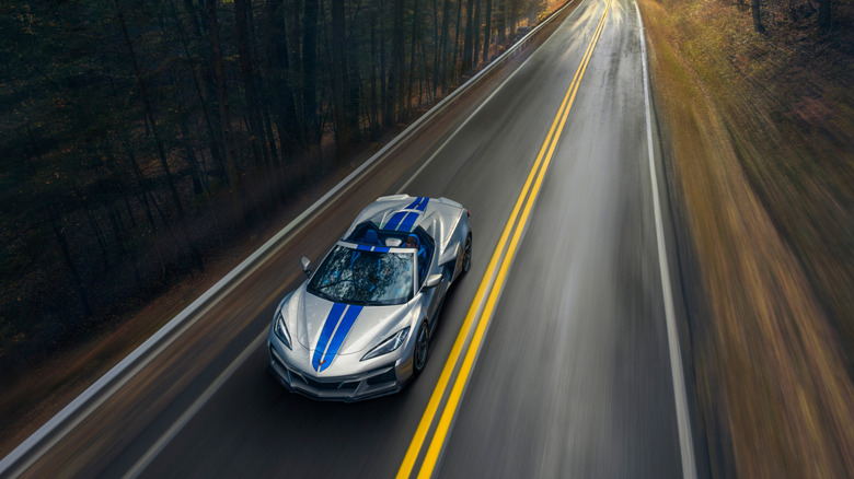 Overhead front view of 2024 Chevrolet Corvette E-Ray 3LZ convertible in Silver Flare with Electric Blue stripe package driving on a road between trees