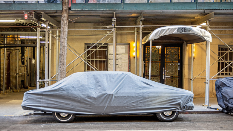 A classic car parked by a sidewalk beneath a car cover