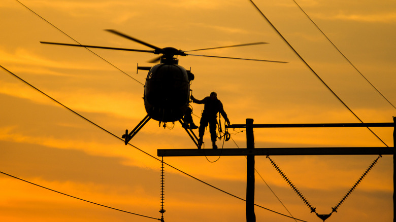 A small helicopter hovers next to a lineman and a series of power lines and telecommunications cables