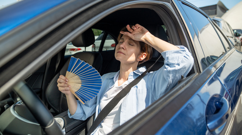 A woman uses a paper fan to cool down while sitting in the driver's seat of a hot car
