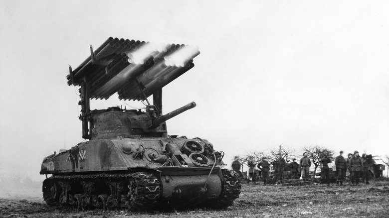 A black and white photo shows an M4 Sherman tank with a Calliope rocket system firing a volley
