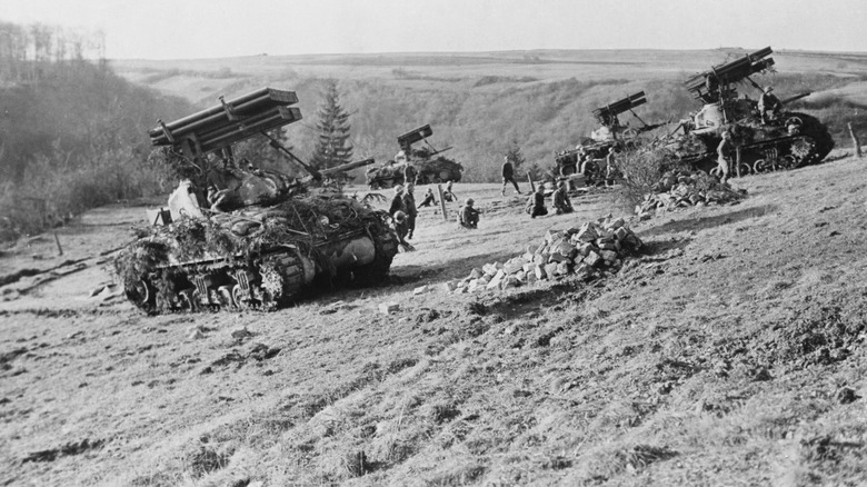 A black and white image shows a series of Sherman tanks advancing up a hill with rocket systems