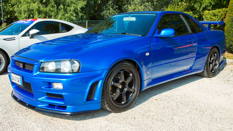 The front 3/4 view of a blue Nissan Skyline GT-R R34 parked on a gravel surface
