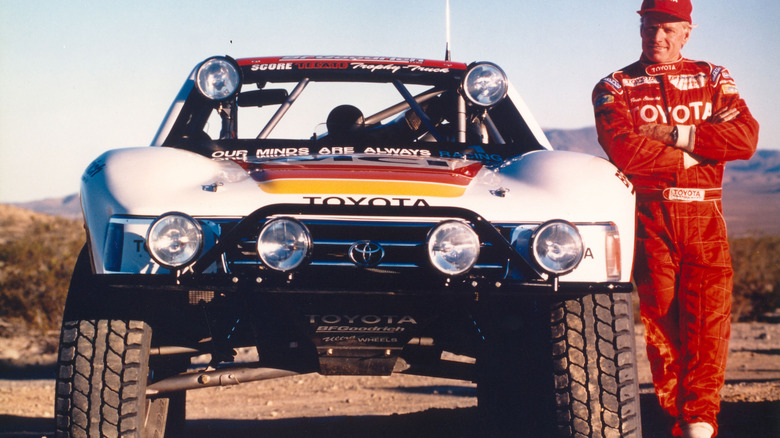 A vintage photo of Ivan 'Ironman' Stewart wearing a red Toyota race suit standing next to his TRD desert race truck parked in the desert