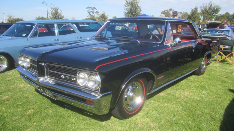 A black 1964 Pontiac GTO parked on grass at a car show with other vehicles in the background