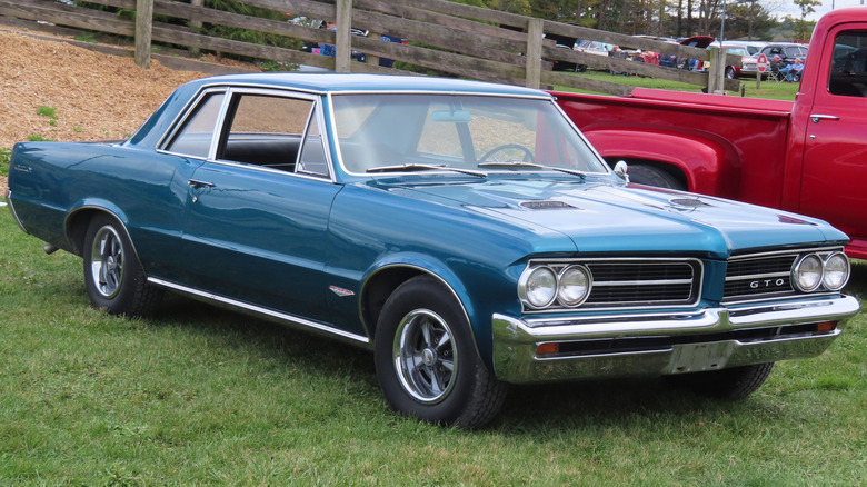 A blue 1964 Pontiac GTO parked on grass at a car show with other vehicles in the background