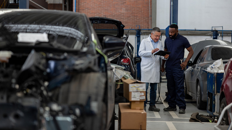 Two mechanics standing in a shop filled with cars in different stages of repair.
