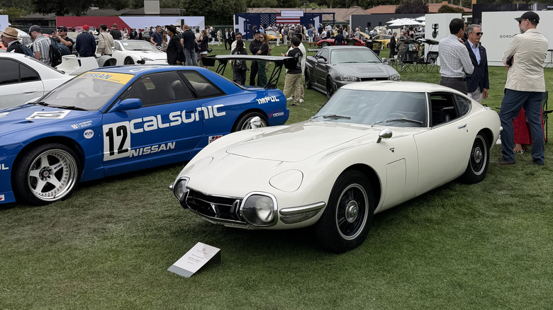 A white Toyota 2000GT parked at the Quail next to a Calsonic Nissan R32 GT-R