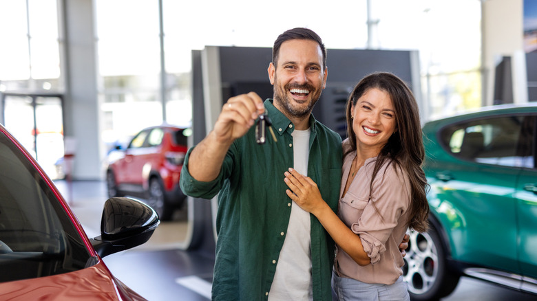 A smiling couple in a car dealership holding up the keys to their new car.