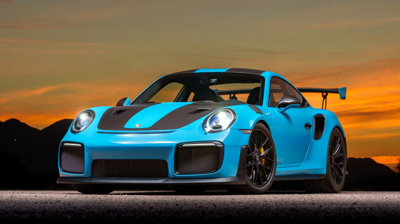 A light blue 2018 Porsche 911 GT2 RS Weissach with its headlights on in front of a sunset and the mountains.