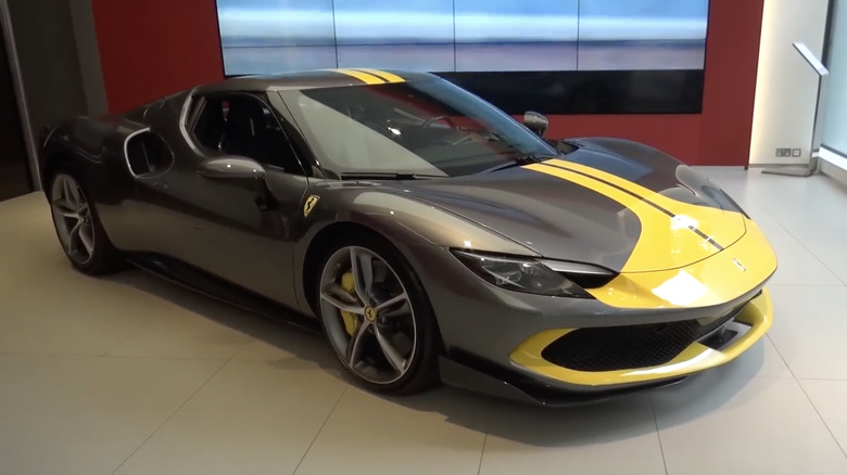 A gray 2023 Ferrari 296 GTB Assetto Fiorano with yellow striping parked in a showroom on a tile floor with a red wall background.