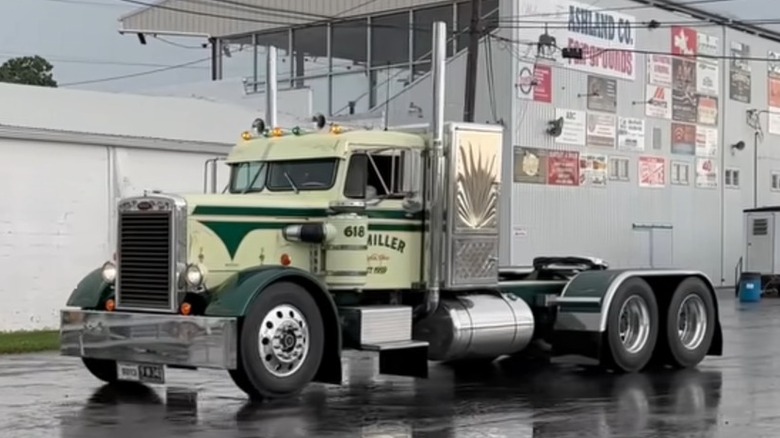 A classic 1950s Peterbilt 351 conventional truck driving on wet asphalt past a low white building. The tractor features a two-tone light cream and dark green paint job, highlighting its signature ultra-narrow "needlenose" butterfly hood layout.