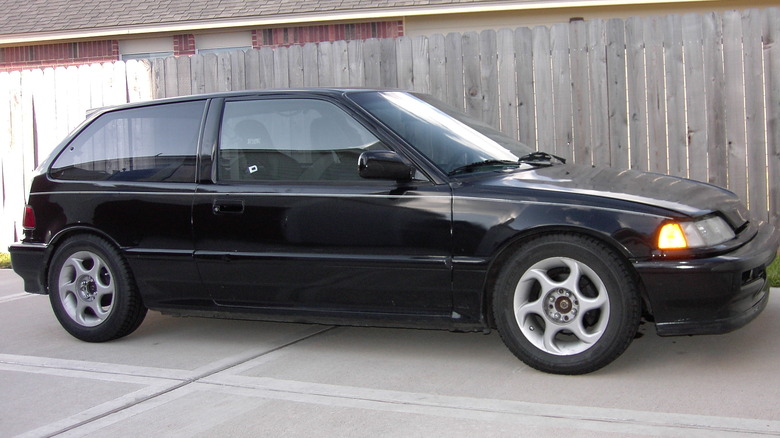 A black 1991 Honda Civic Si parked on a concrete driveway in front of a wooden fence and brick house