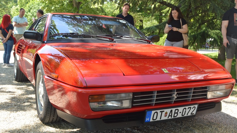 A red Ferrari Mondial T coupe sitting on a gravel road with trees and onlookers behind it