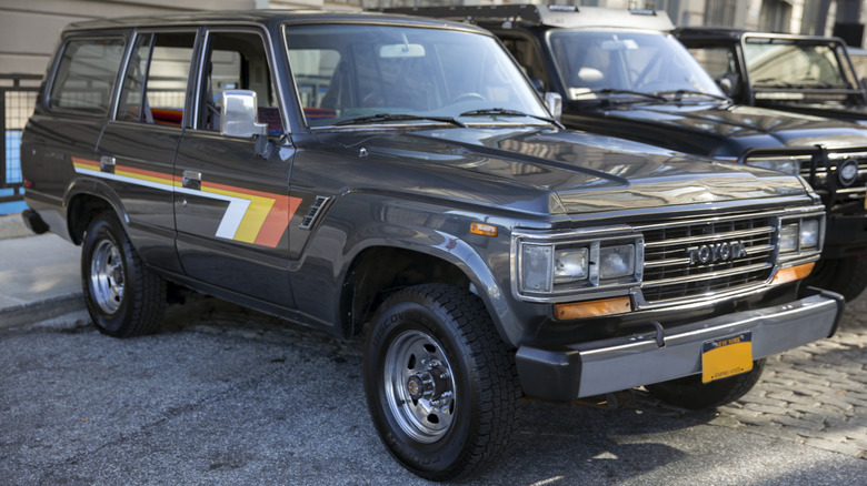 A gray 1990 Toyota Land Cruiser FJ62 with white, yellow, and orange stripes parked in a lot in front of a fence, next to other off-road vehicles