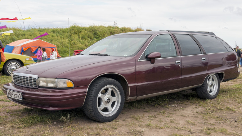 A burgundy Chevrolet Caprice Classic station wagon sits in a field in front of bushes at a car show