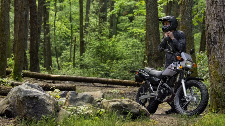 A rider standing next to a 2026 Yamaha TW200 in a forest surrounded with green trees