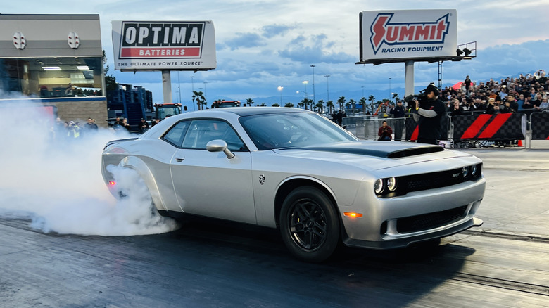 A silver Dodge Demon 170 in doing a burnout at the dragstrip with a crowd and signs for Summit Racing and Optima Batteries in the background