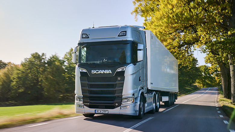 A wide, low-angle dynamic shot of a modern, silver Scania 770S cab-over semi-truck traveling down a two-lane paved rural highway.