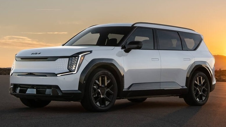 A white Kia EV9 electric SUV parked on a wide, open asphalt tarmac during a dramatic sunset.