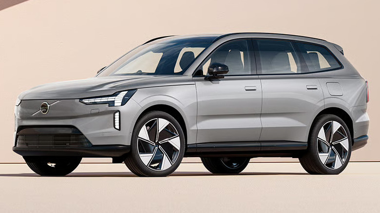 A silver Volvo EX90 luxury electric SUV parked on a clean, sand-colored surface against a minimalist architectural backdrop
