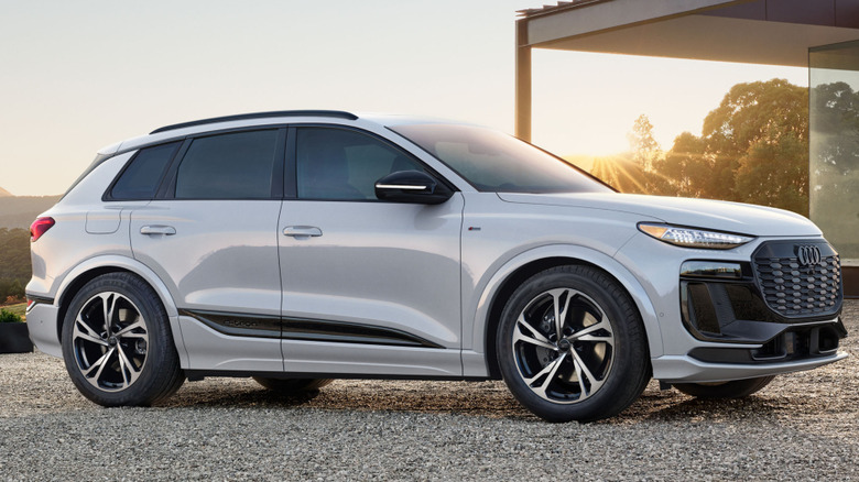 A white Audi Q6 e-tron electric SUV parked on a wide, textured gravel courtyard, positioned next to a contemporary single-story home.