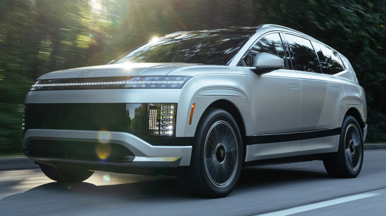 A silver Hyundai Ioniq 9 electric SUV driving down a paved road with the sun shining in the background.