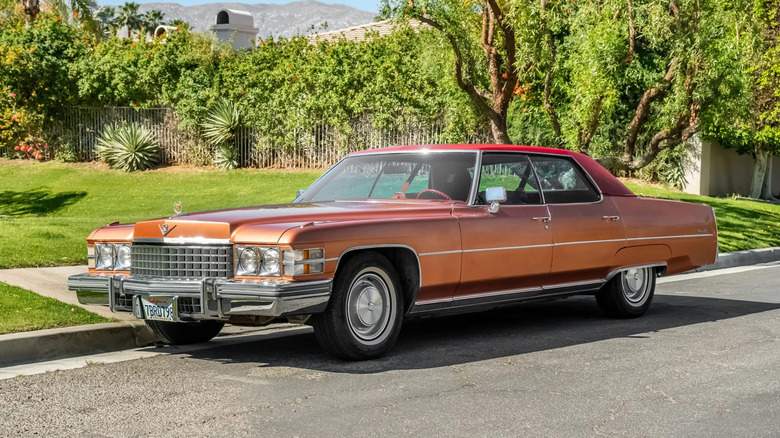 Front three quarters shot of a brown Cadillac Sedan DeVille parked on the street in front of a grassy yard