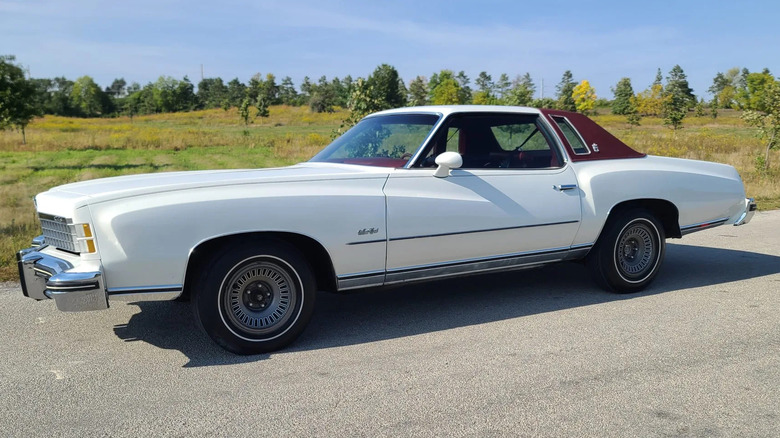 Side view of a white Monte Carlo with a burgundy Landau roof parked in front of a field of grass and trees