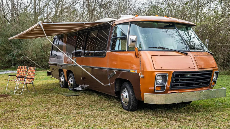 An orange and brown GMC Motorhome parked on grass with its striped awning open and two matching chairs outside it