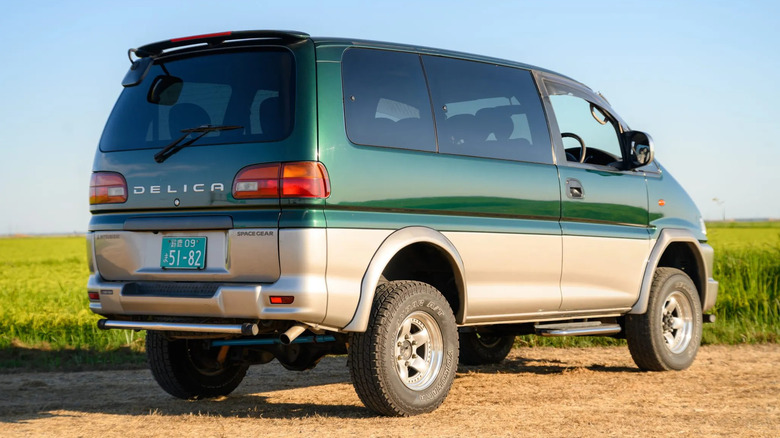 rear three-quarters shot of a green Mitsubishi Delica Space Gear parked on dirt in front of a green field
