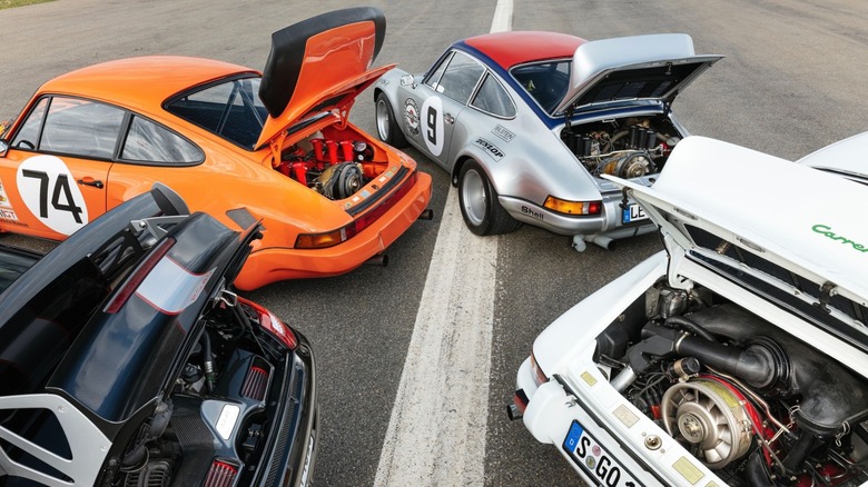 An overhead group shot of four classic air-cooled Porsche 911 models parked on a track with all of their rear engine hoods raised to display various generations of the iconic Hans Mezger-designed flat-six engine and large axial cooling fans.