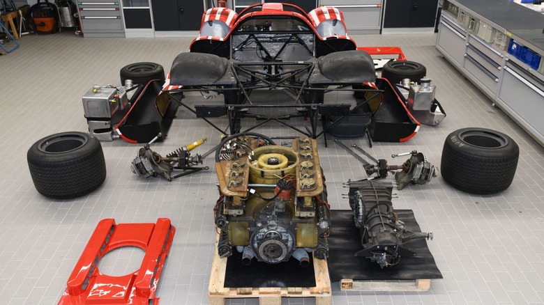 A workspace floor showing a disassembled Porsche 917 race car with its red-and-white body shell in the background, while the massive Type 912 flat-12 engine with its iconic yellow horizontal cooling fan sits on a wooden pallet in the foreground