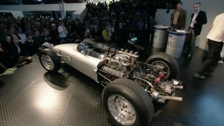 A silver Porsche 804 open-wheel Formula 1 race car displayed on a stage before a seated crowd, with its rear bodywork completely removed to reveal the intricate, high-revving Type 753 flat-8 racing engine