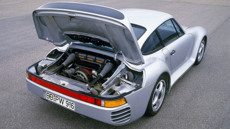 A rear three-quarter view of a silver Porsche 959 parked on an asphalt track with its large rear engine decklid fully raised, exposing the highly complex, hybrid air-and-water-cooled twin-turbocharged flat-six engine.