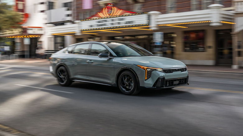 A side view of a teal Kia K4 driving past a blurry theater in an urban setting