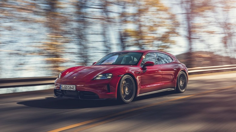 A Carmine Red Porsche Taycan GTS Sport Turismo wagon driving at speed along a tree-lined road, front three-quarter panning shot.