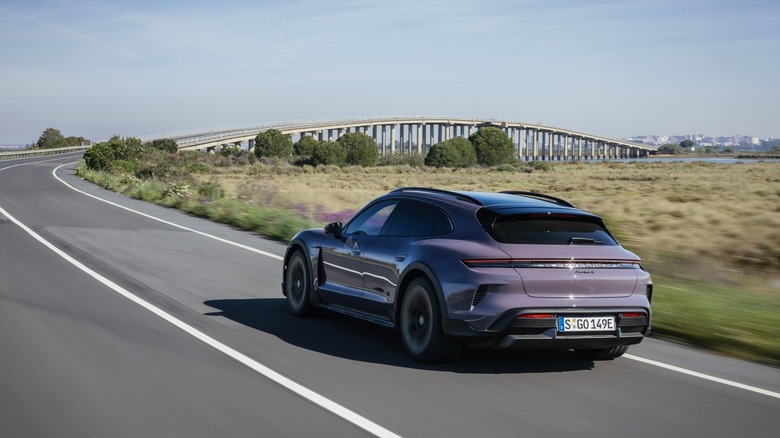 A Porsche Taycan Turbo S Cross Turismo wagon in pale purple driving down an open coastal road toward a bridge, rear three-quarter view.