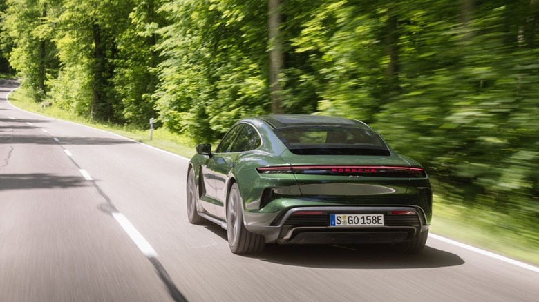A forest green Porsche Taycan Turbo sedan driving along a paved road through a lush green forest, rear three-quarter view.
