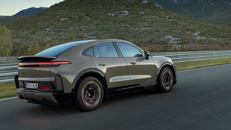 A Porsche Cayenne Turbo Electric Coupe in olive metallic driving on a racetrack with hills in the background, rear three-quarter view.